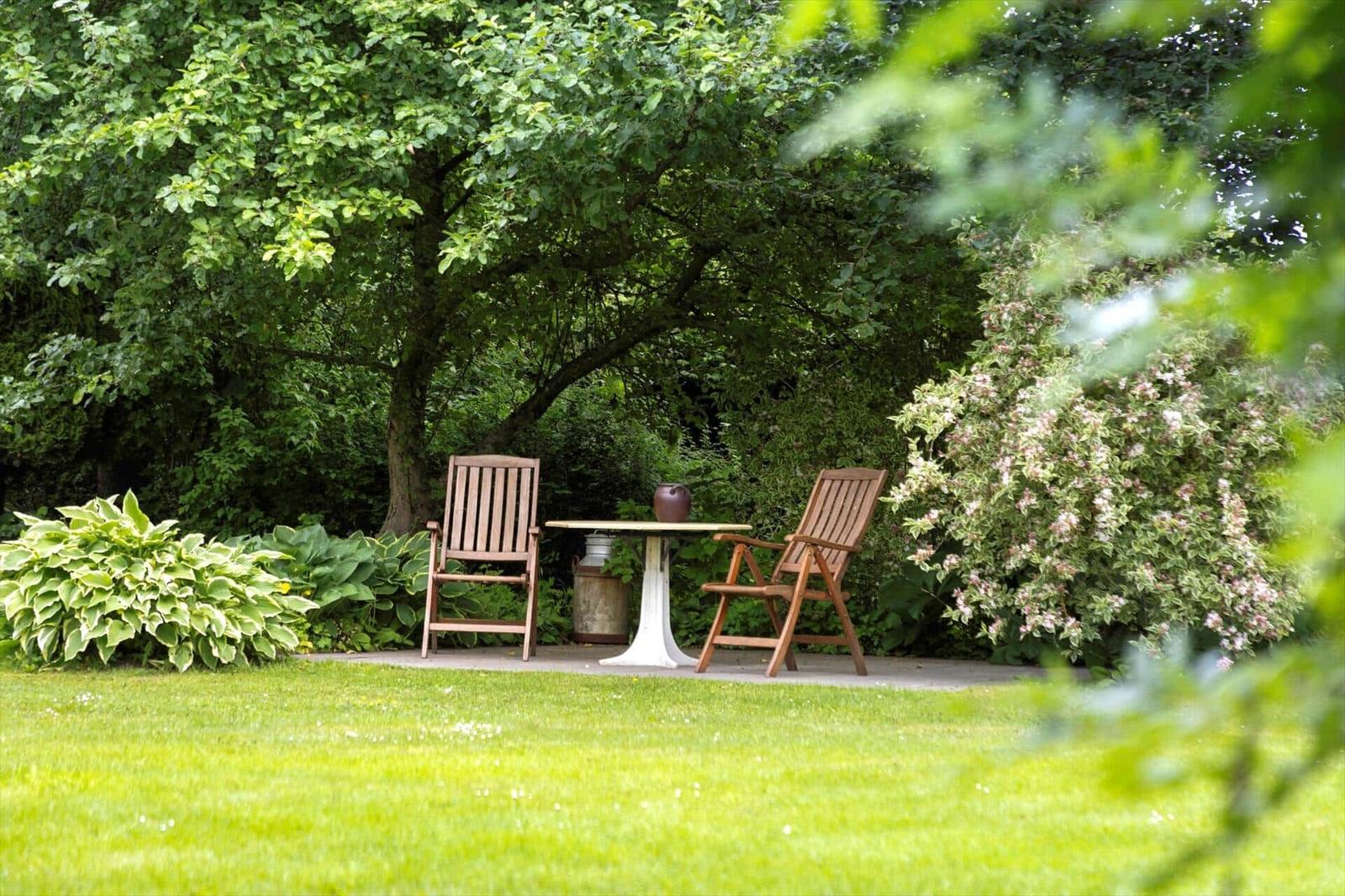 Zwei Holzstühle und ein Tisch stehen auf einer Terrasse im Garten unter Bäumen.