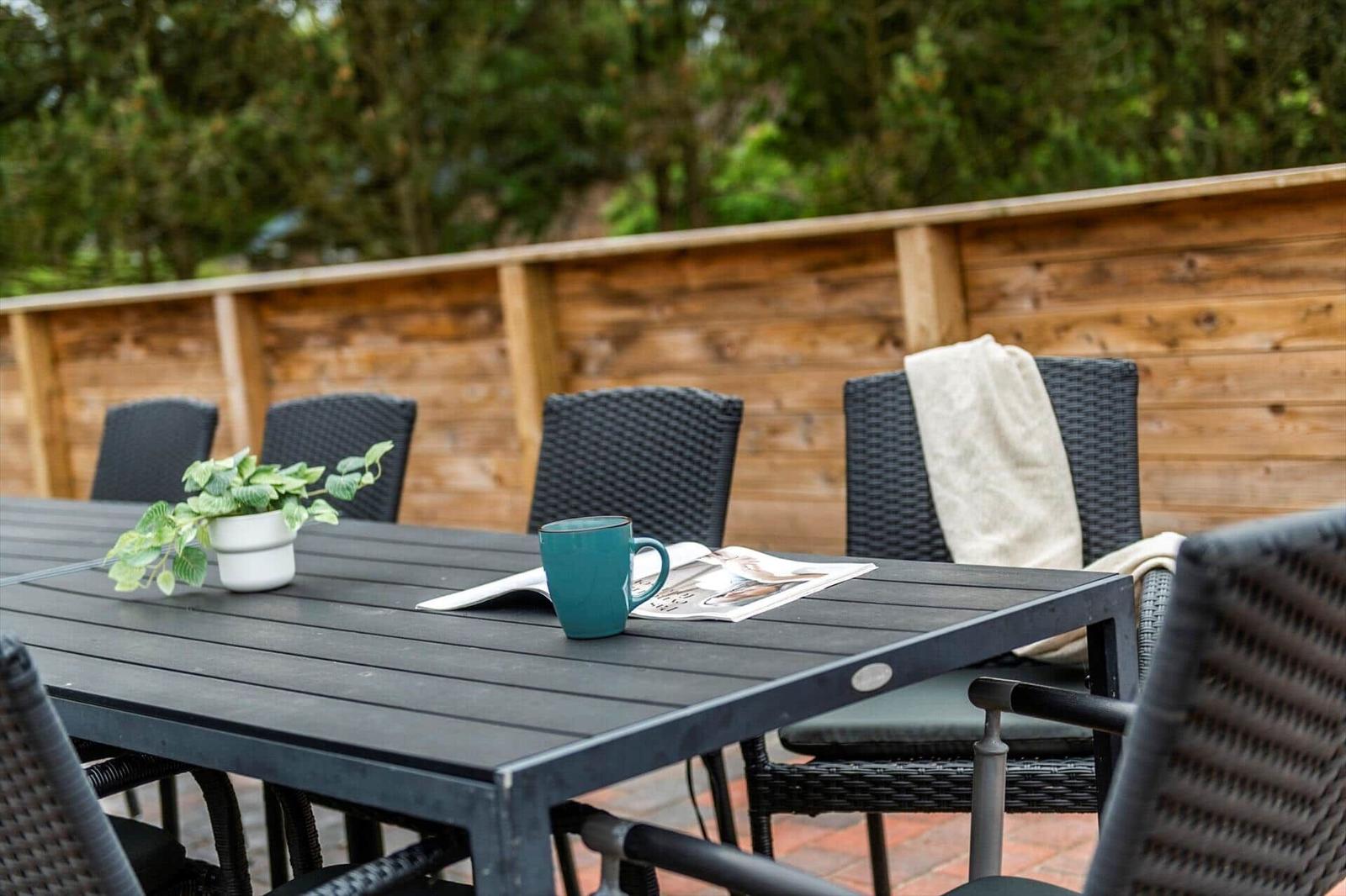 Terrasse mit Tisch, Stühlen, Pflanze und Tasse. Hintergrund: Holzgeländer und Bäume.