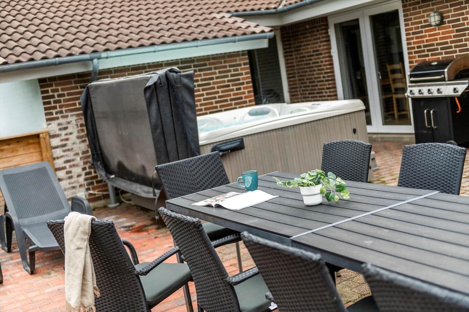 Terrasse mit Tisch, Stühlen, Whirlpool und Grill. Hintergrund: Backsteinhaus mit Dach.