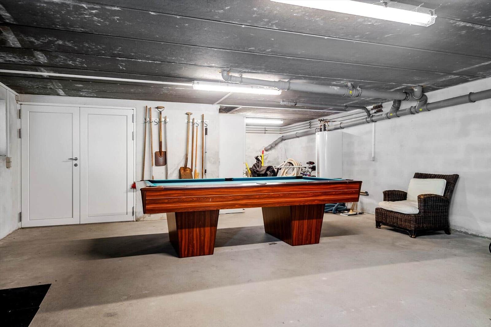 Pool table with blue felt and wooden frame in basement. Tools hang on the wall. A seating area stands beside it.