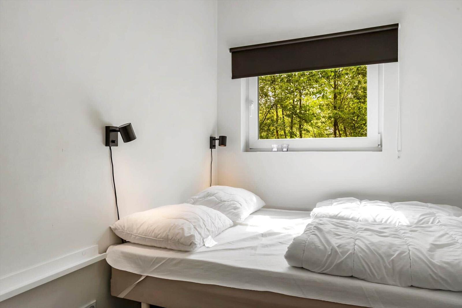 Schlafzimmer mit weißem Bett und Fensterblick auf Wald.