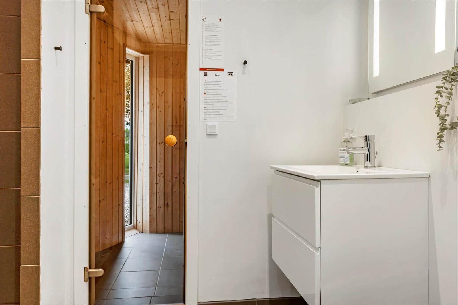 Badezimmer mit Waschbecken und Spiegel. Tür führt in Sauna mit Holzwand.