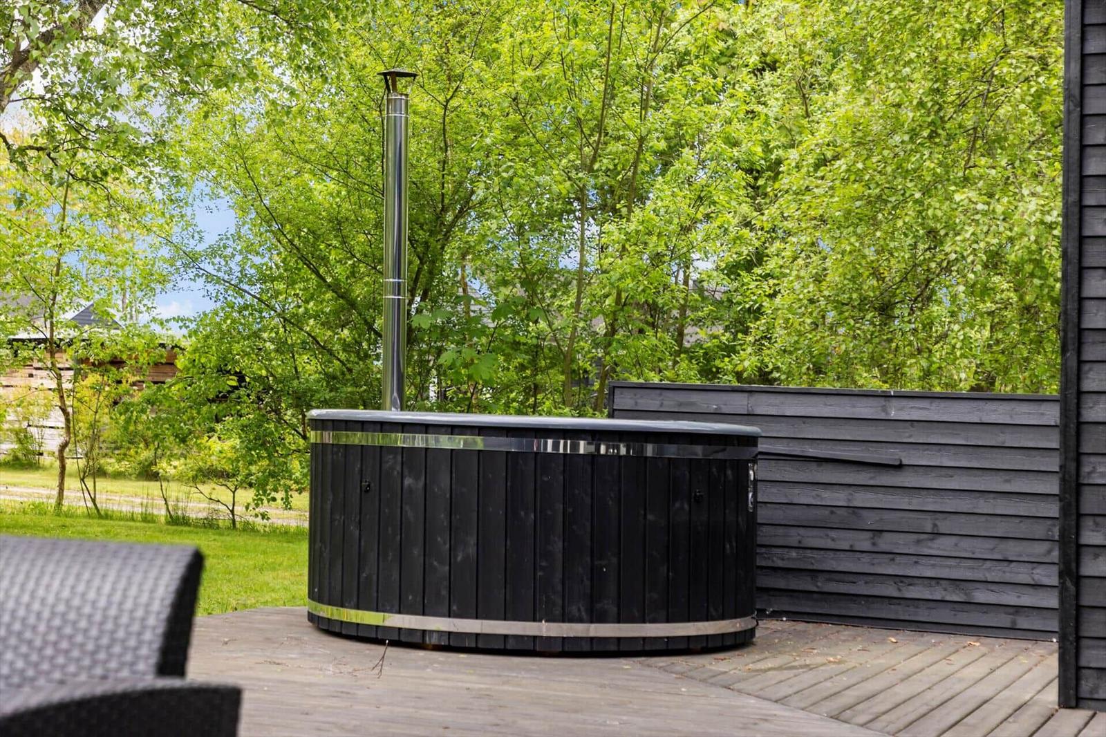 Schwarzer Whirlpool auf Holzterrasse mit grüner Umgebung und Metallrohr.