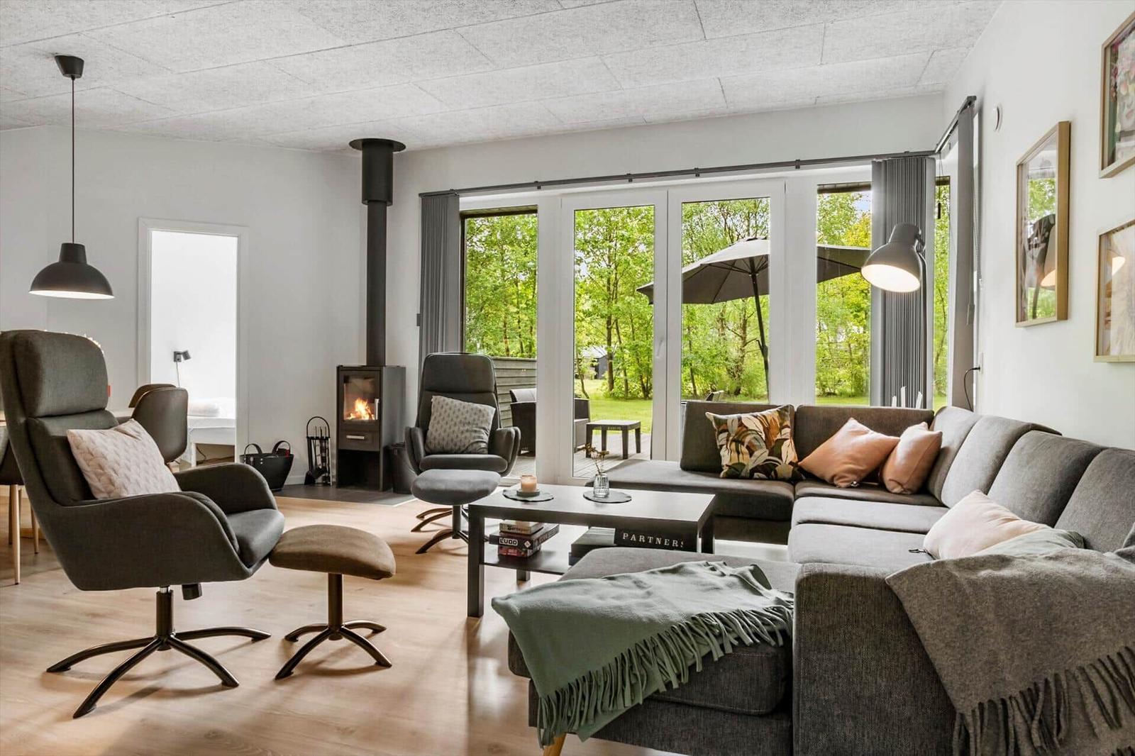 Wohnzimmer mit Sofa, Sesseln und Kamin. Durchgangstür zur Terrasse mit Blick auf Grünfläche.