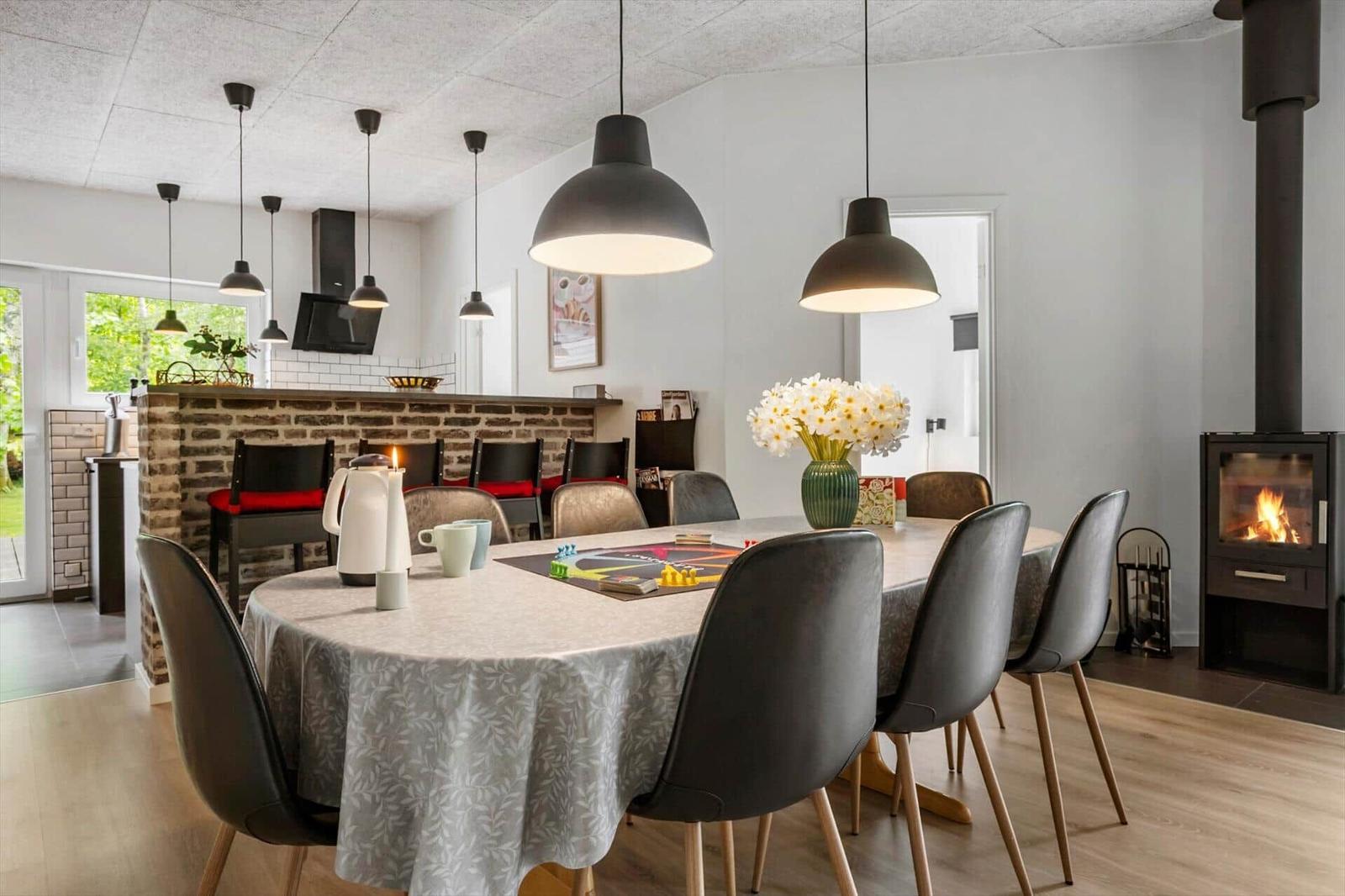 Esszimmer mit runder Tisch, schwarzen Stühlen und Kaminofen. Hängelampen und Blumen auf Tisch.