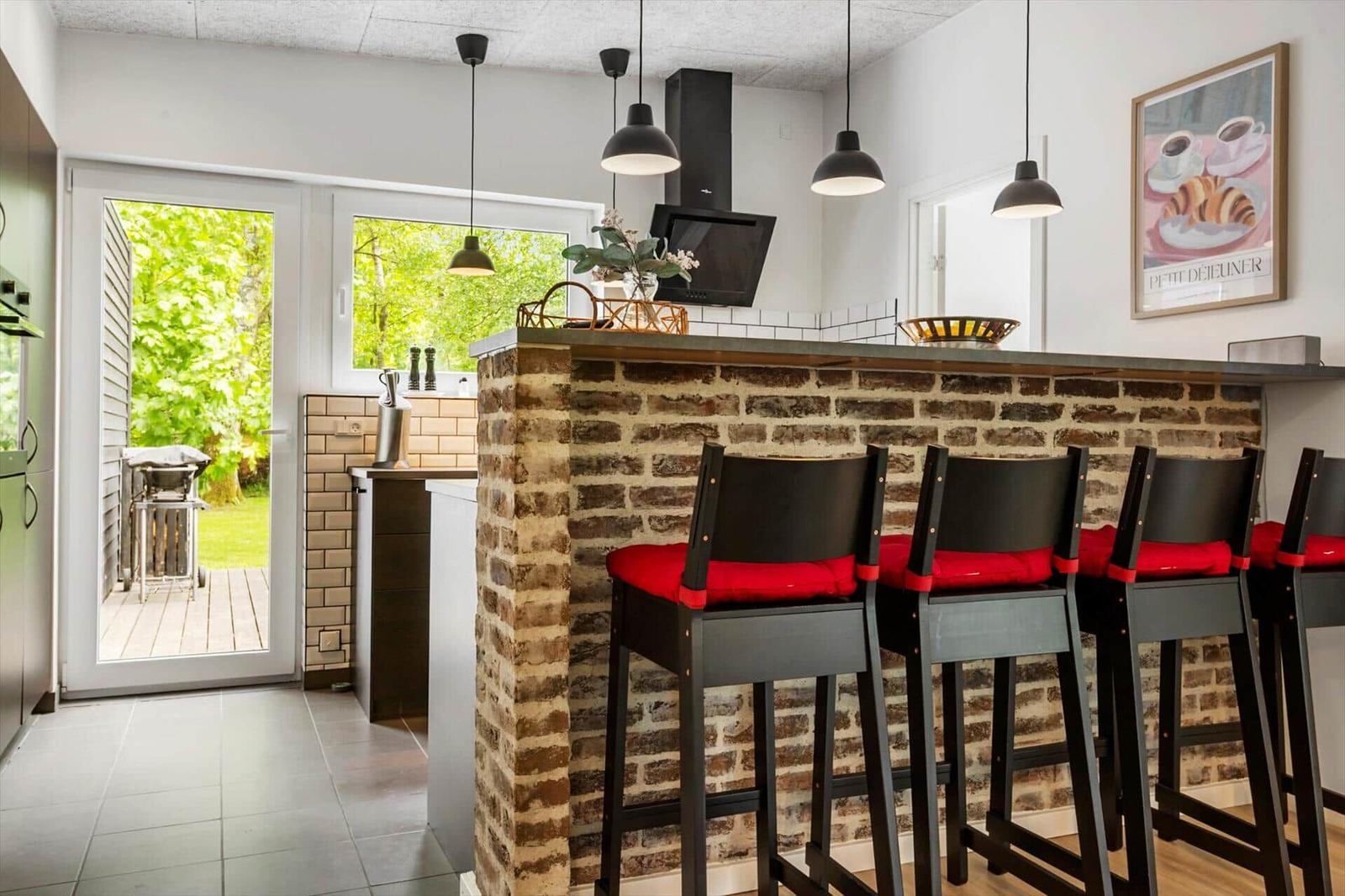 Küchenbereich mit Steinbar und roten Sitzkissen. Durchgang zur Terrasse mit Blick auf Grün.