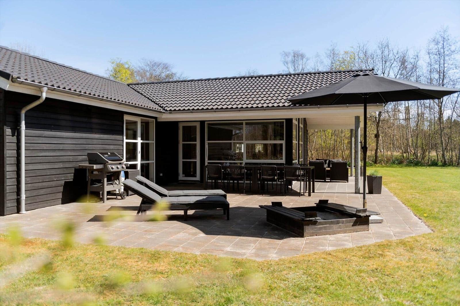Terrasse mit Grill, Liegen und Feuerkorb vor einem Haus mit dunkler Fassade.
