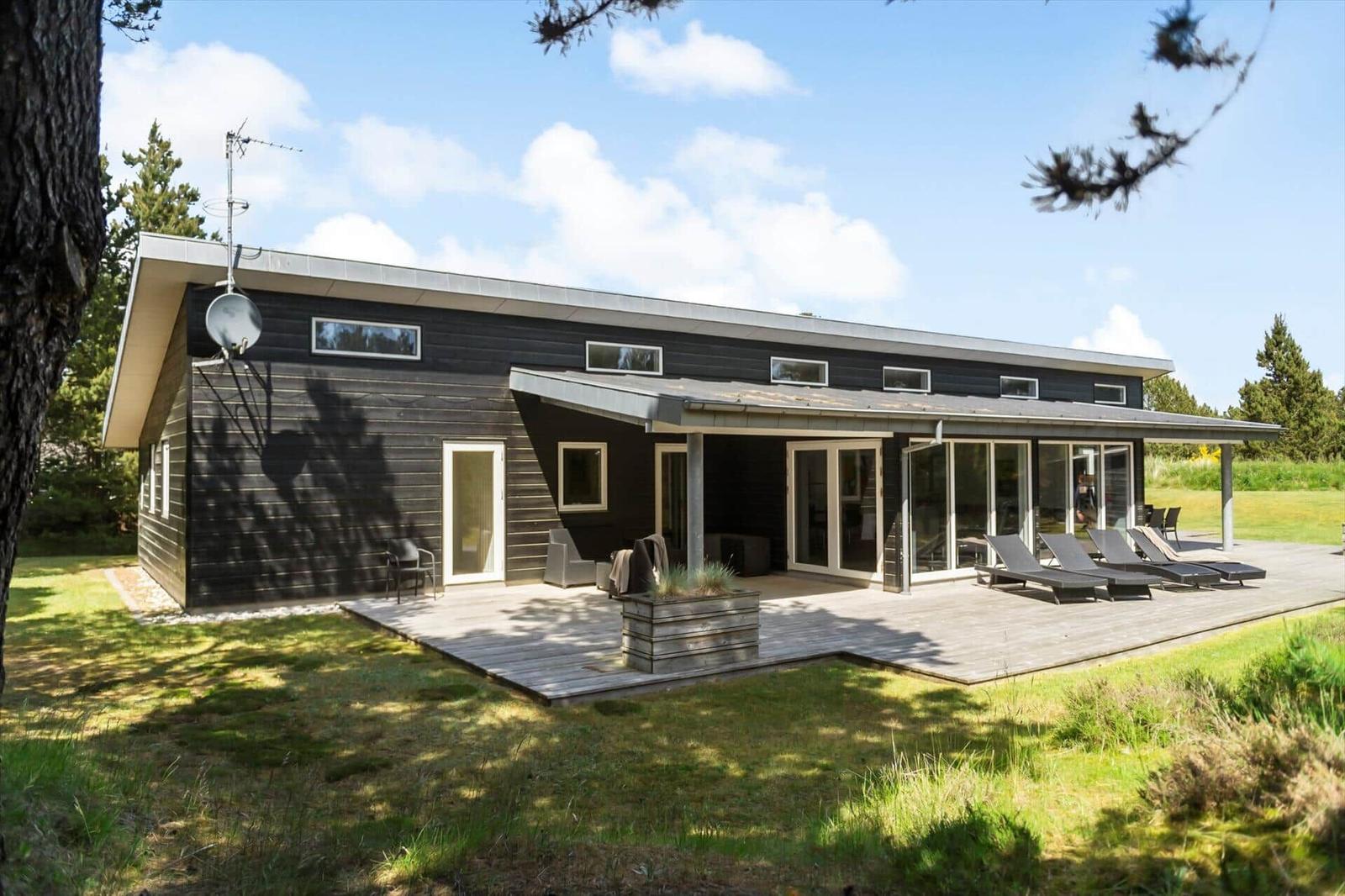 Moderne Holzhütte mit Terrasse, Sonnenliegen und Garten. Satellitenschüssel und große Fenster sichtbar.