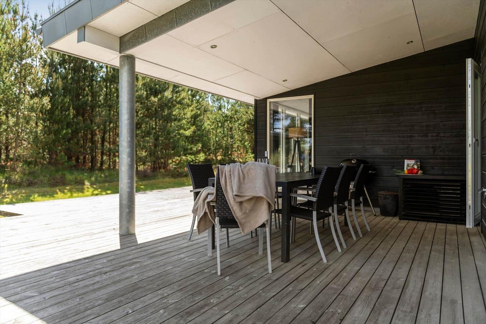 Terrasse mit Tisch, Stühlen und Grill. Hintergrund: Wald. Holzdecke und dunkle Wand.