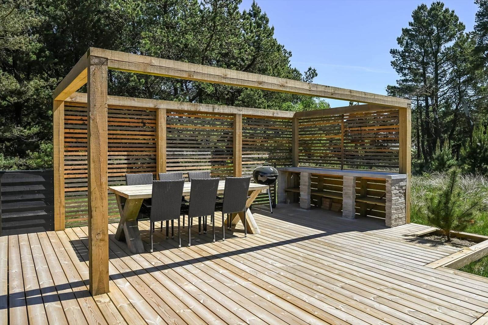 Holzterrasse mit Pavillon, Tisch, Stühlen und Grill. Umgeben von Bäumen und Gras.