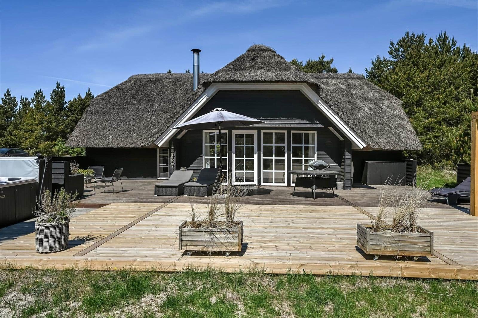 Haus mit Strohdach und großem Holzterrasse mit Gartenmöbeln und Grillplatz.