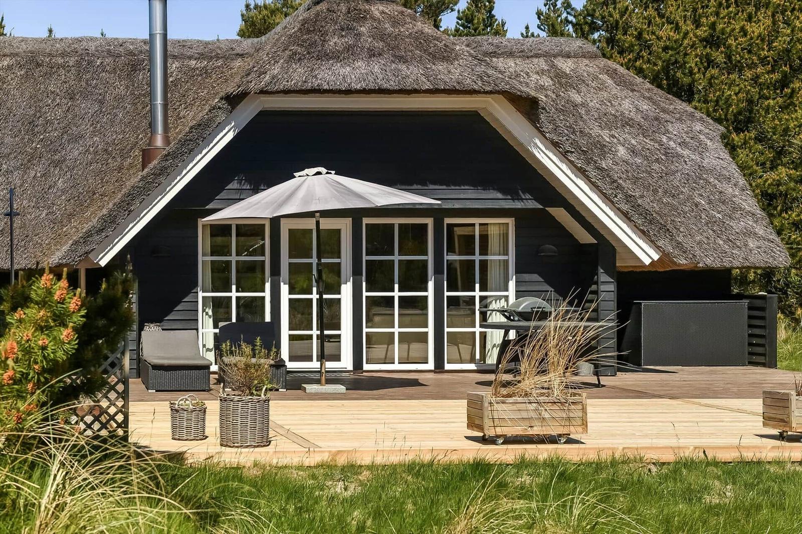 Haus mit Strohdach und Holzterrasse mit Gartenmöbeln und Sonnenschirm.