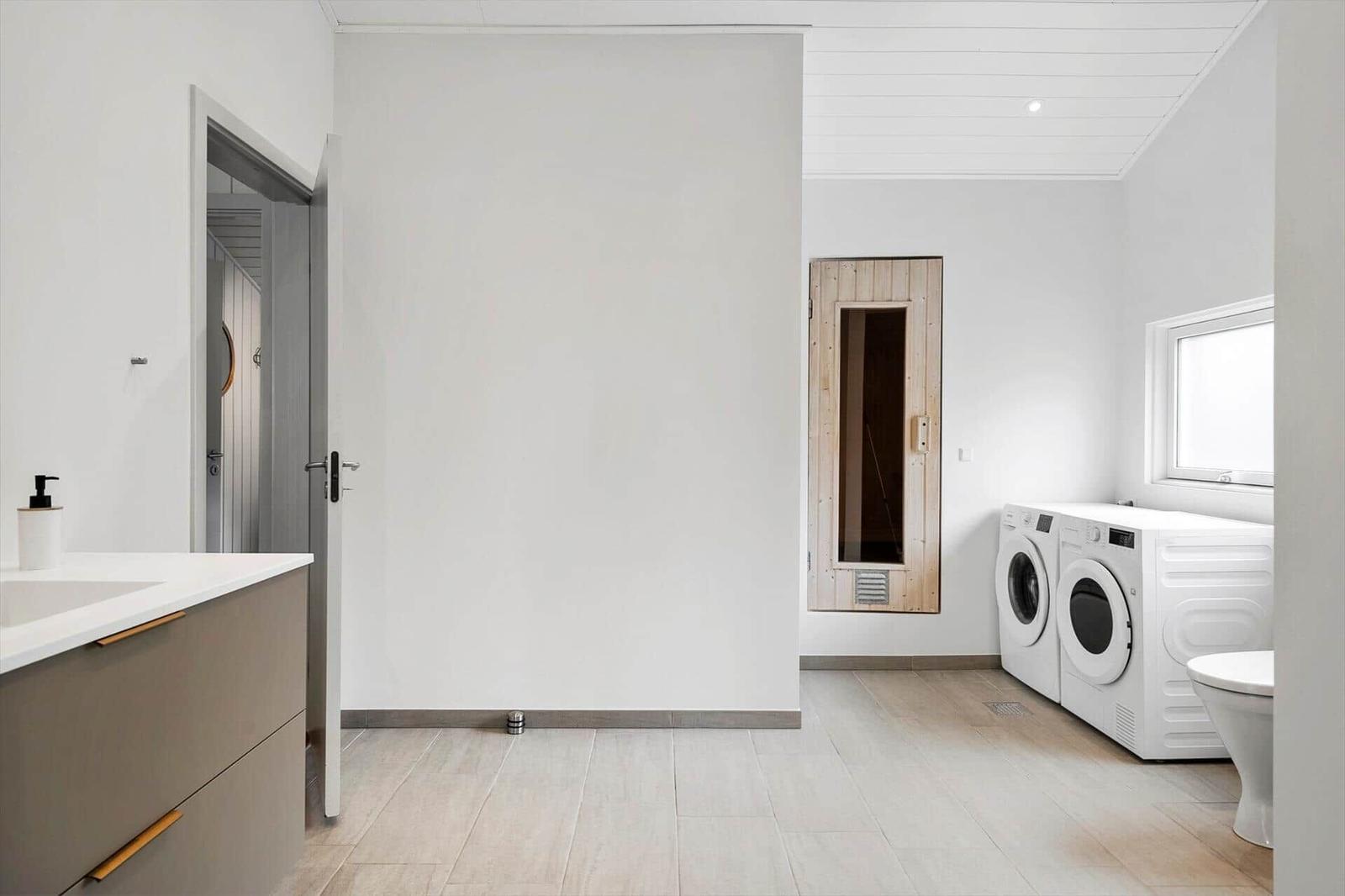 Waschraum mit Waschmaschine, Trockner und Toilette. Holztür führt in Sauna.