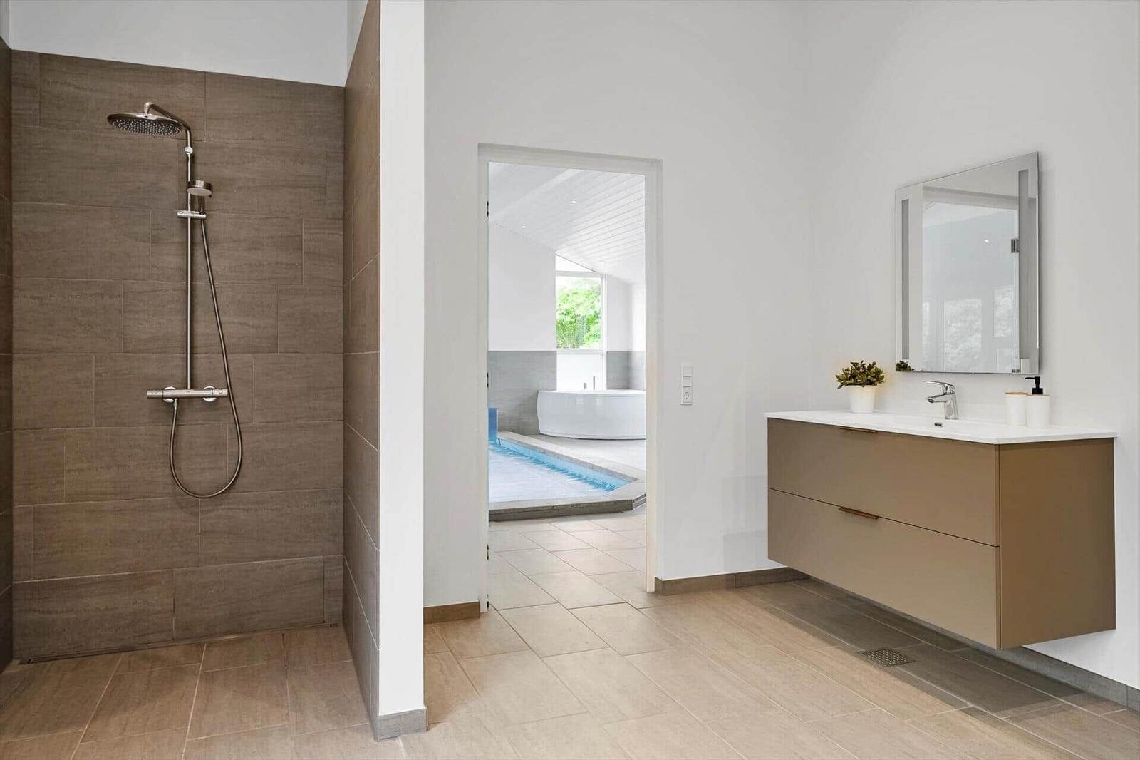 Dusche mit Duschbrause, Waschbecken und Spiegel. Durchgang zur Badewanne und Pool.