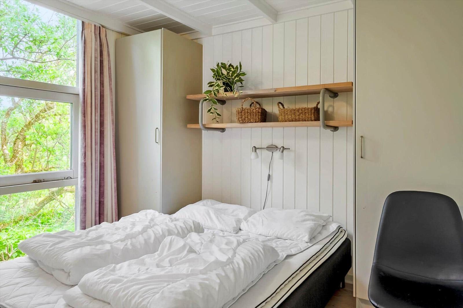 Schlafzimmer mit Bett, Fenster und Wandregalen. Draußen sind Bäume sichtbar.