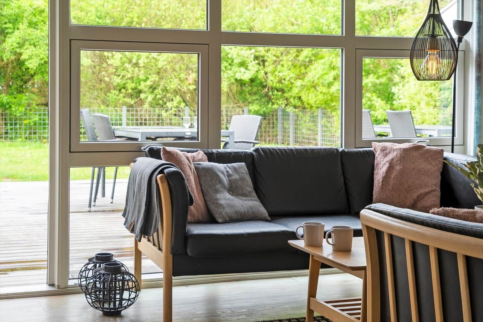 Ein Wohnbereich mit Sofa, Tisch und Sitzgelegenheiten. Durch große Fenster ist eine Terrasse mit Garten sichtbar.