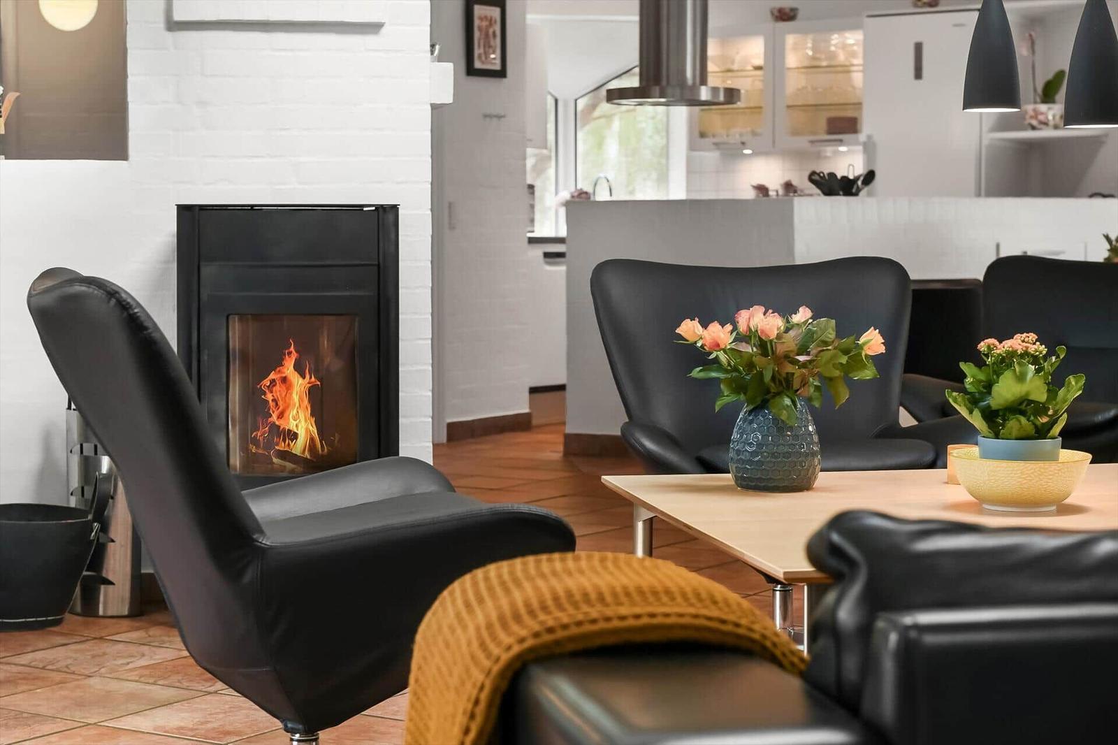 Wohnbereich mit Kamin, schwarzen Sesseln und Holztisch mit Blumen.