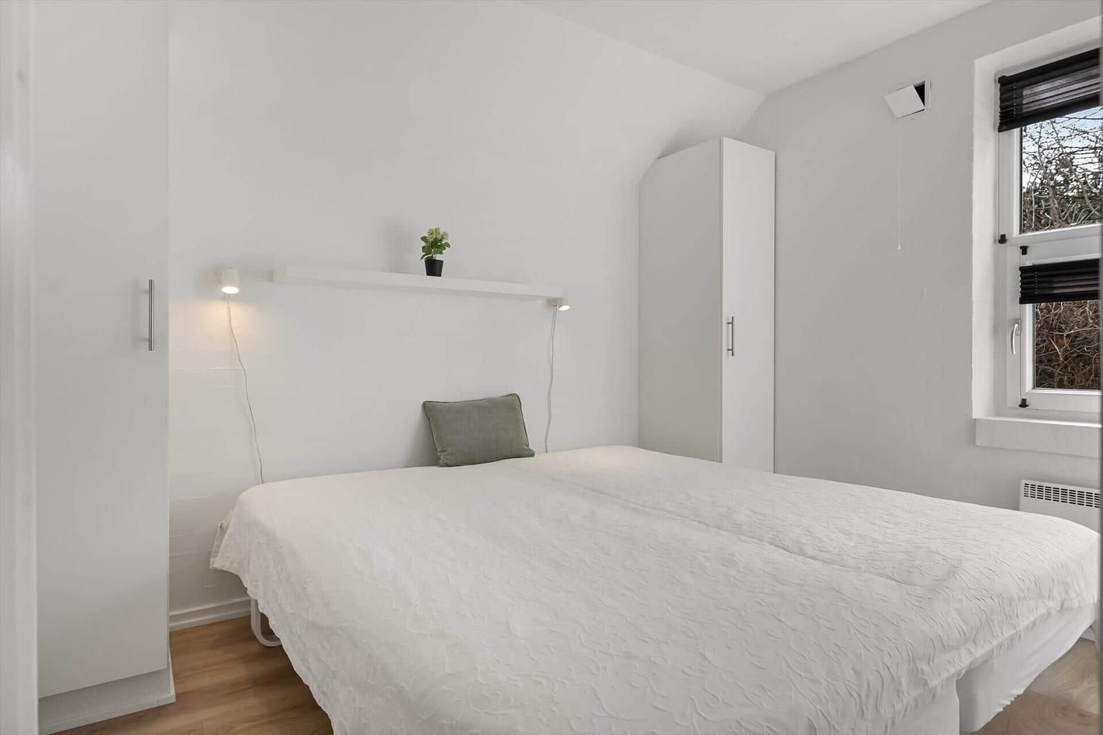 Schlafzimmer mit weißem Bett, Kleiderschränken und Fenster mit Jalousie.