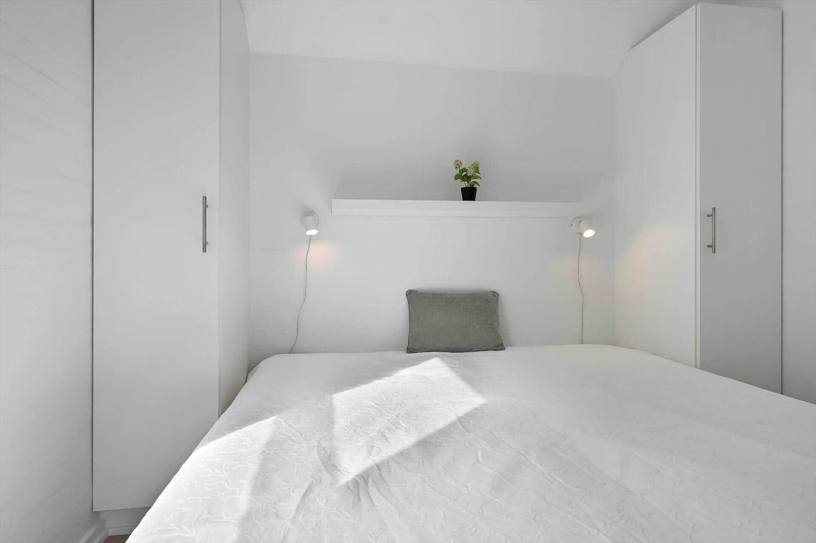Schlafzimmer mit weißem Bett, grauem Kissen und weißen Schränken. Auf Regal eine Pflanze und zwei Wandlampen.