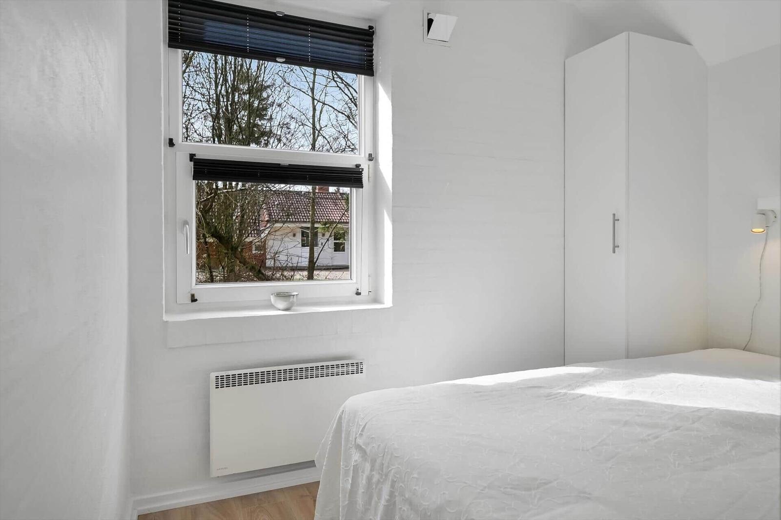 Schlafzimmer mit weißem Bett, Kleiderschrank und Fenster mit Blick auf Außenbereich.