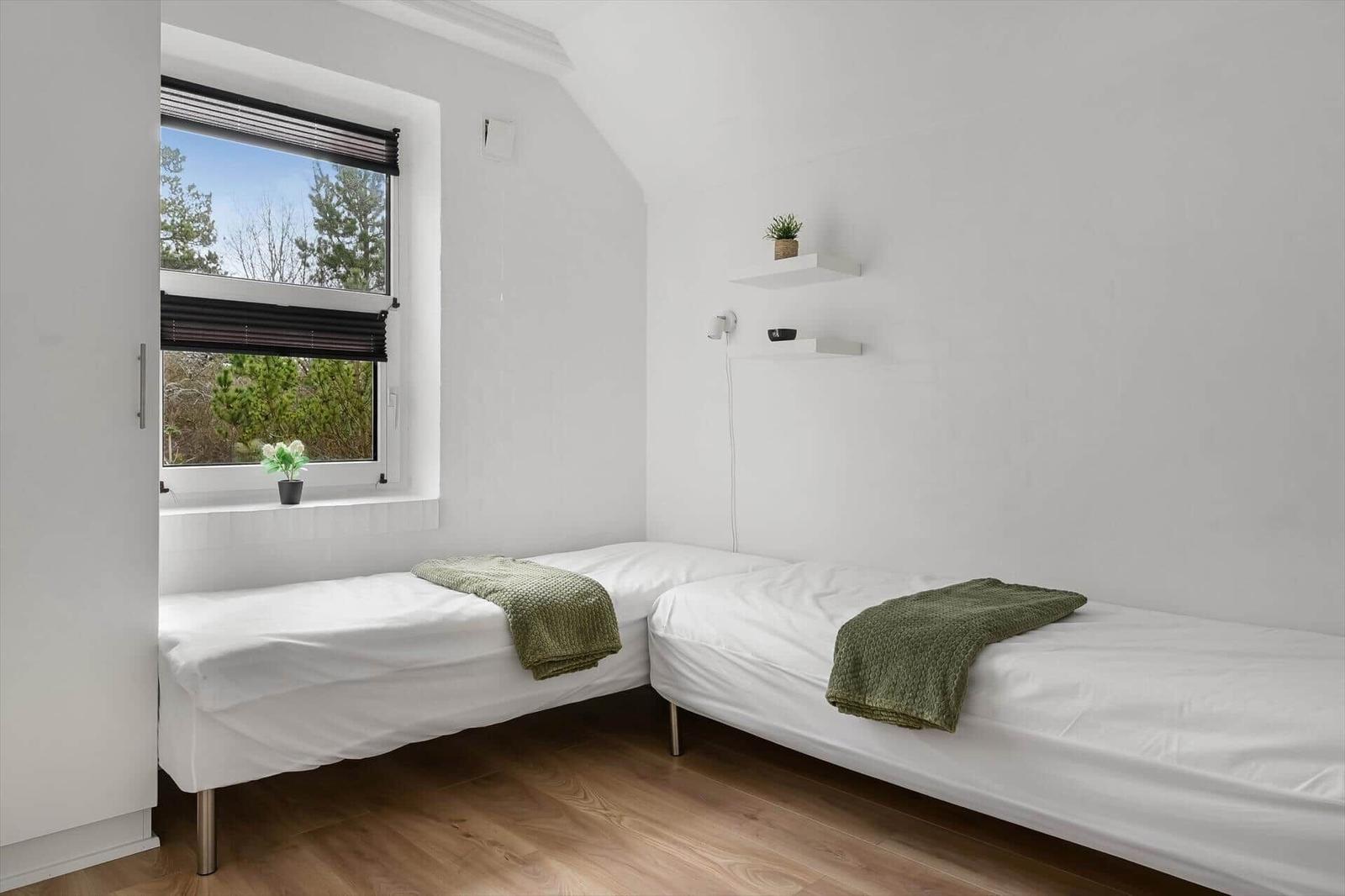 Zwei Einzelbetten mit grünen Decken. Fenster mit Blick auf Bäume. Holzboden und weiße Wände.