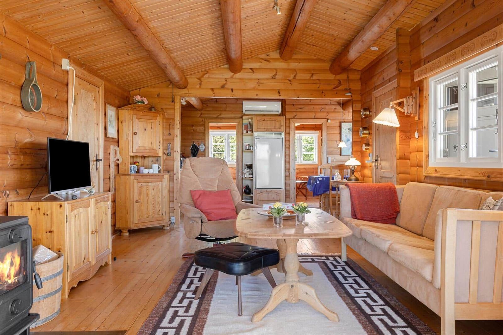 Wohnzimmer mit Holzdecke, Sofa, Sessel und Esstisch. Kamin und Küchenbereich sichtbar.
