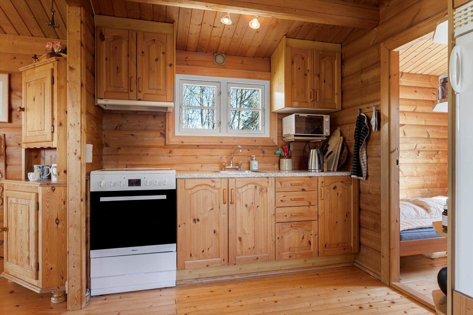 Küche mit Holzmöbeln, Herd, Spüle und Fenster. Blick in Schlafzimmer mit Bett.