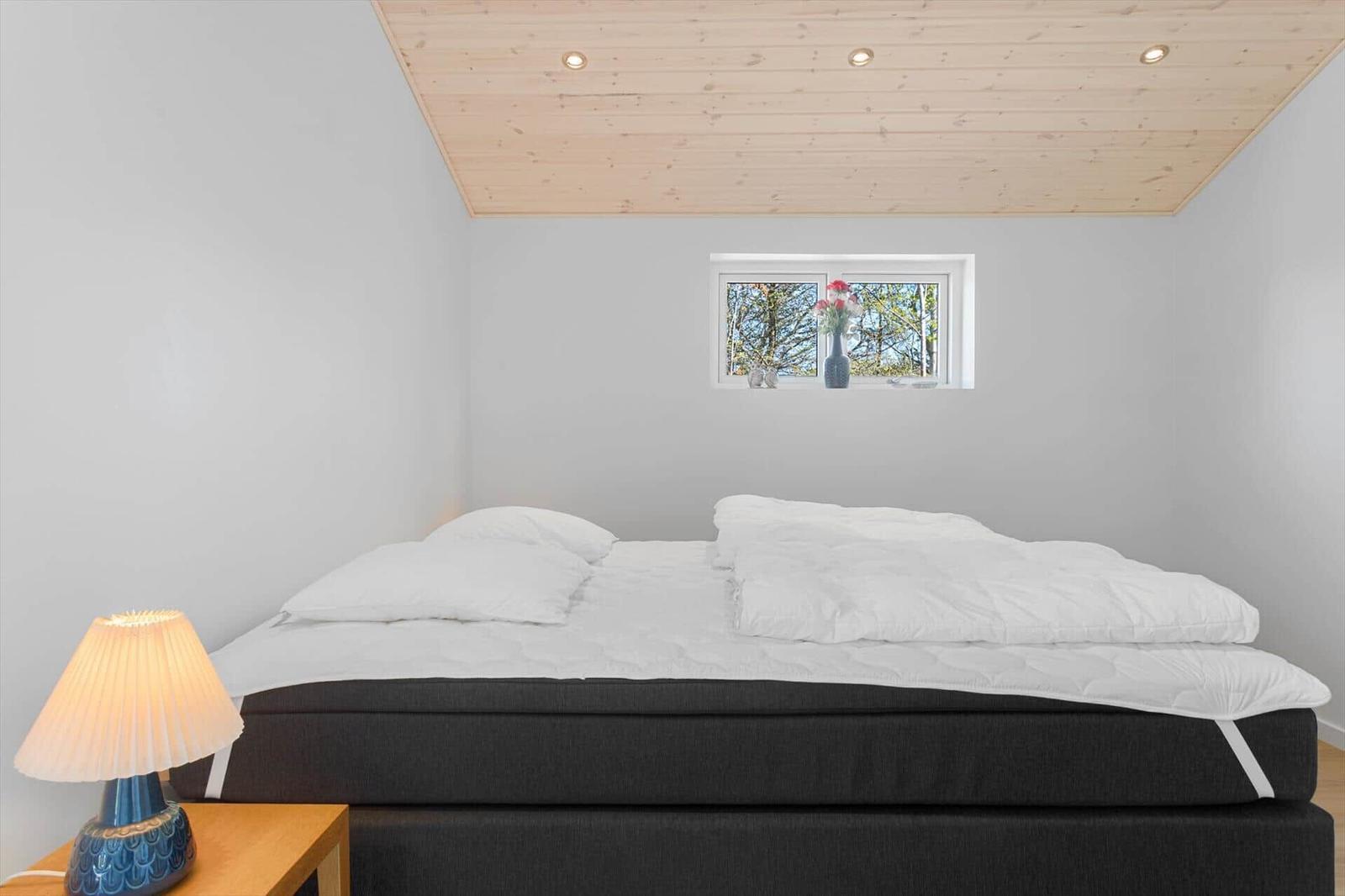 Schlafzimmer mit Bett, Nachttisch und Lampe. Holzdecke und kleines Fenster mit Blick auf Bäume.