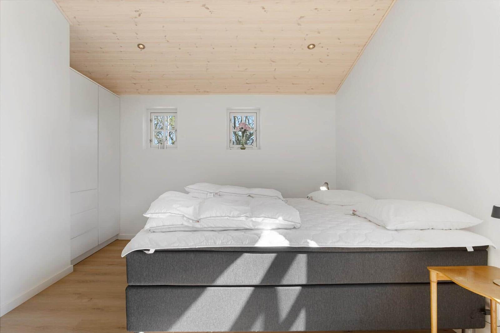 Schlafzimmer mit Bett, Kleiderschrank und zwei Fenstern.