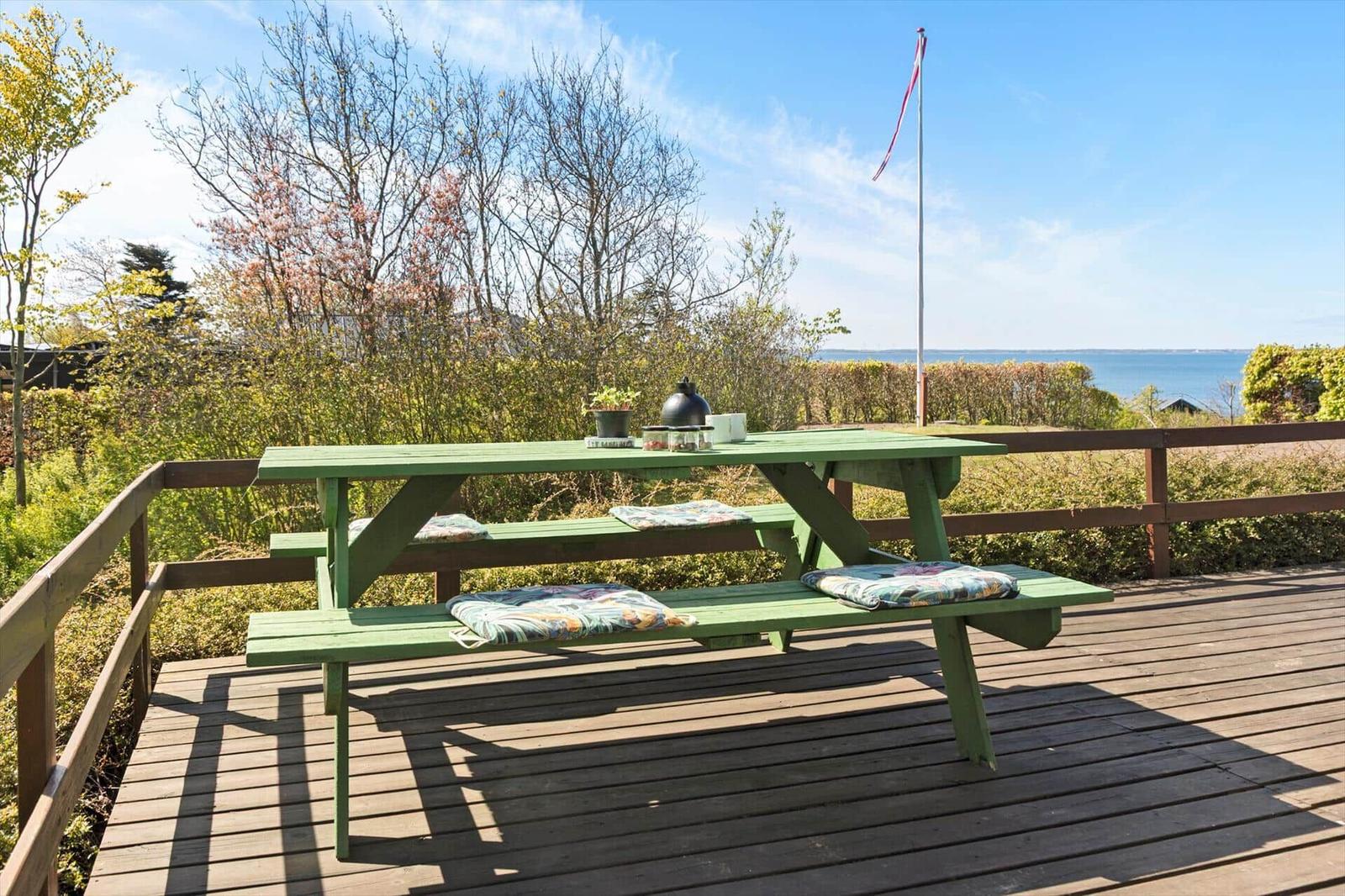 Grüner Picknicktisch mit Kissen auf Terrasse mit Blick auf Wasser und Bäume.