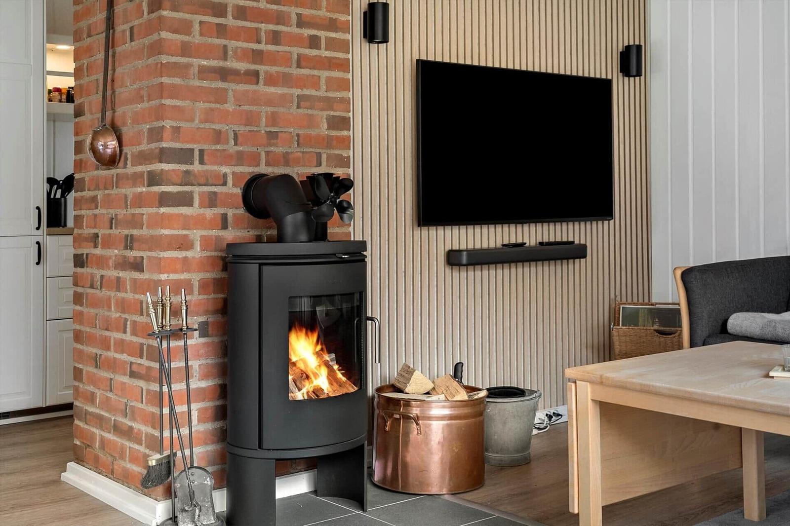 Ein Kaminofen mit brennendem Holz steht neben einem Fernseher an einer Holzwand.