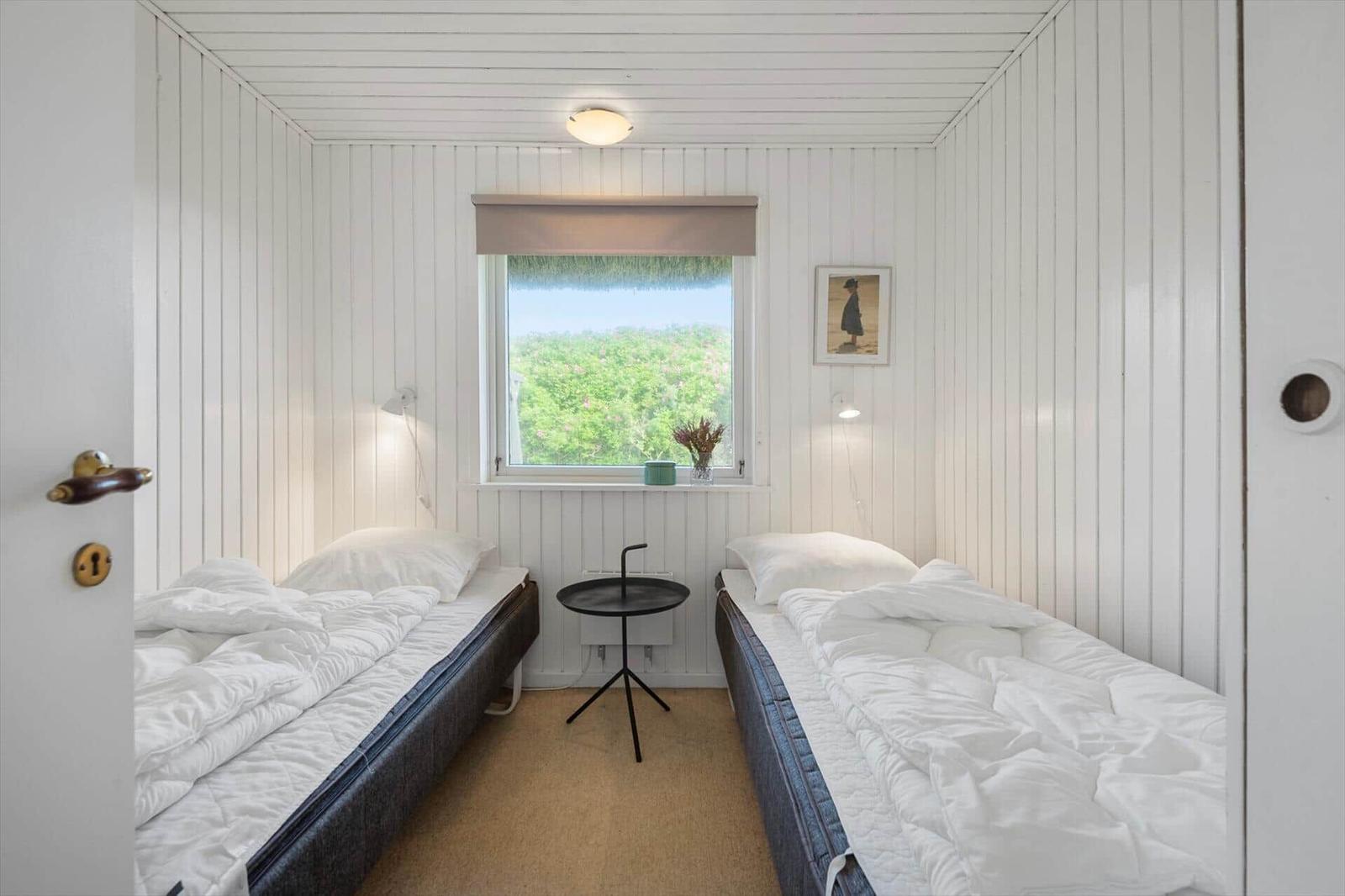 Two beds, window with view of greenery, small table and wall picture.