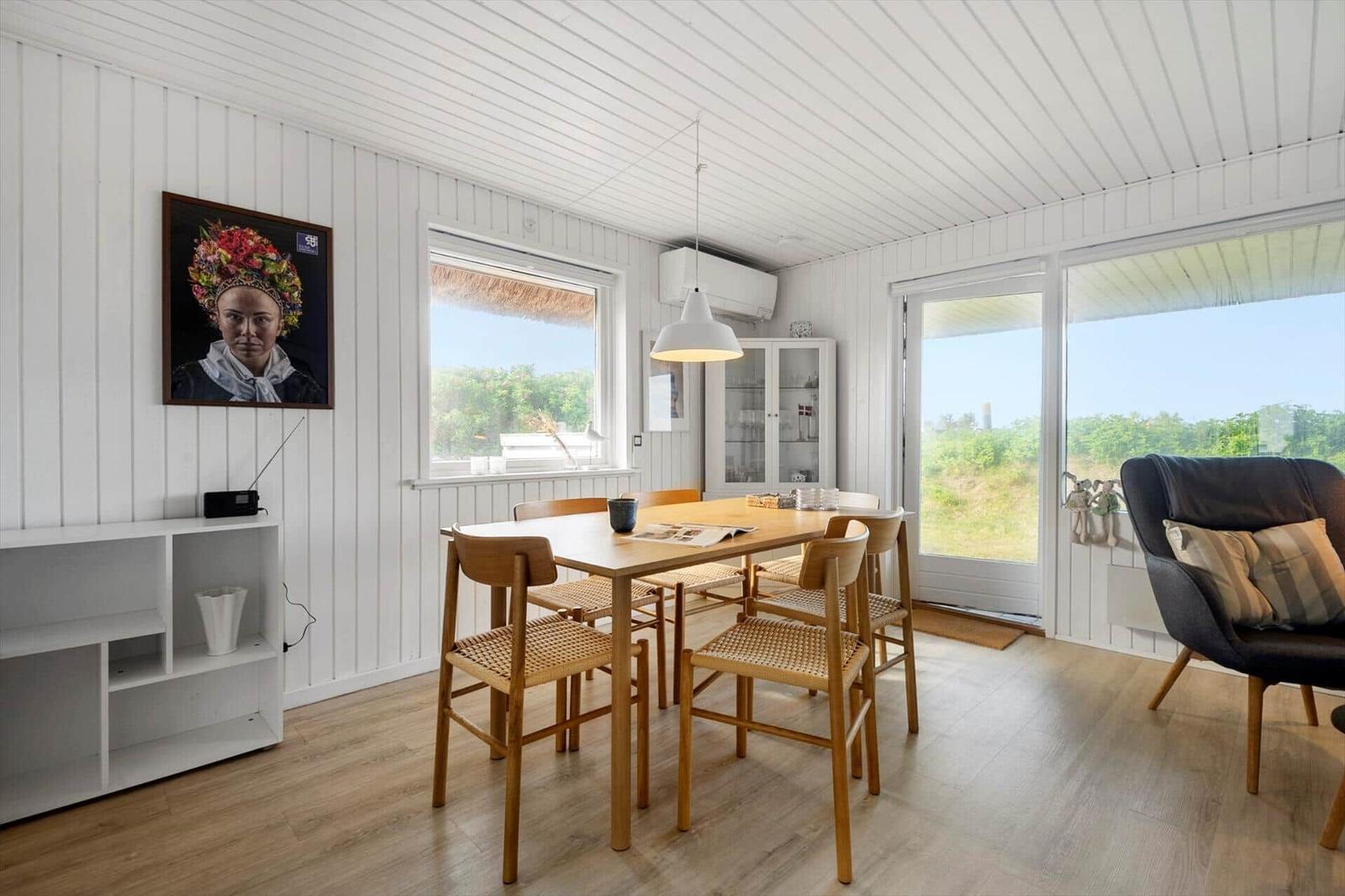 Dining room with wooden table, chairs, and large windows. Wall art and cabinet are visible.