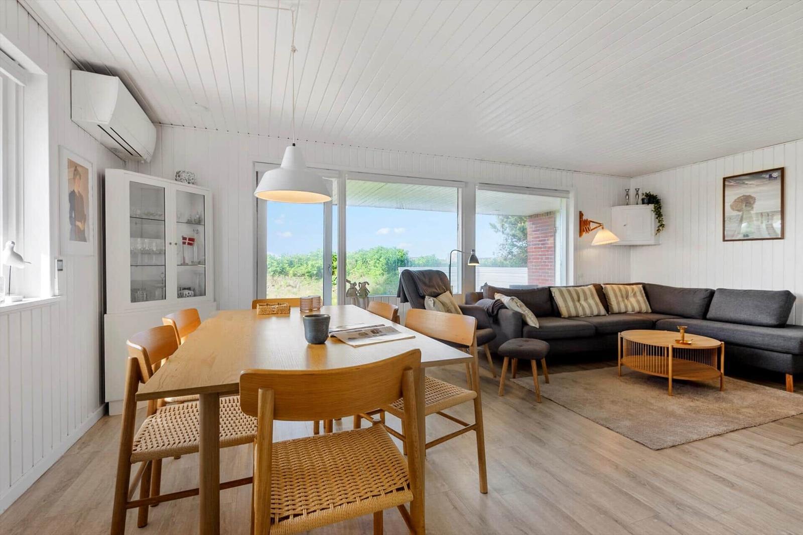 Dining area and living room with wooden table, chairs, and sofa. Large windows with view of the landscape.