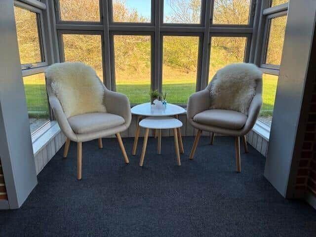 Zwei Sessel mit Fellbezügen stehen um einen runden Tisch herum. Große Fenster bieten Blick auf Grünflächen.
