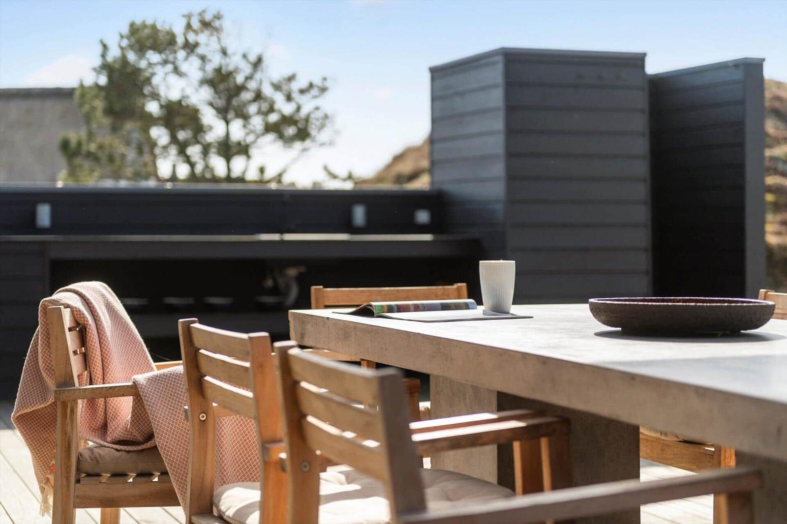 Holztisch mit Stühlen auf Terrasse. Hintergrund: moderne Hauswand und Baum.