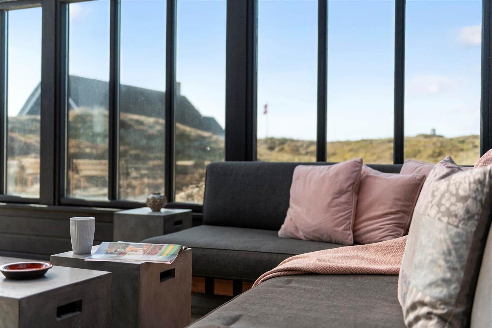 Ein Wohnbereich mit grauem Sofa, rosa Kissen und großem Fensterblick auf Landschaft.
