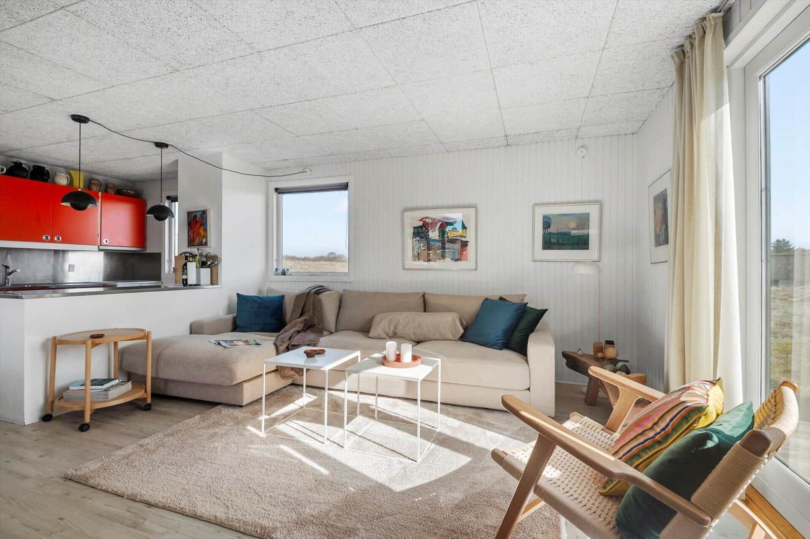 Wohnbereich mit Sofa, Sessel und Küchenzeile. Große Fenster, Wandbilder und rote Schränke.