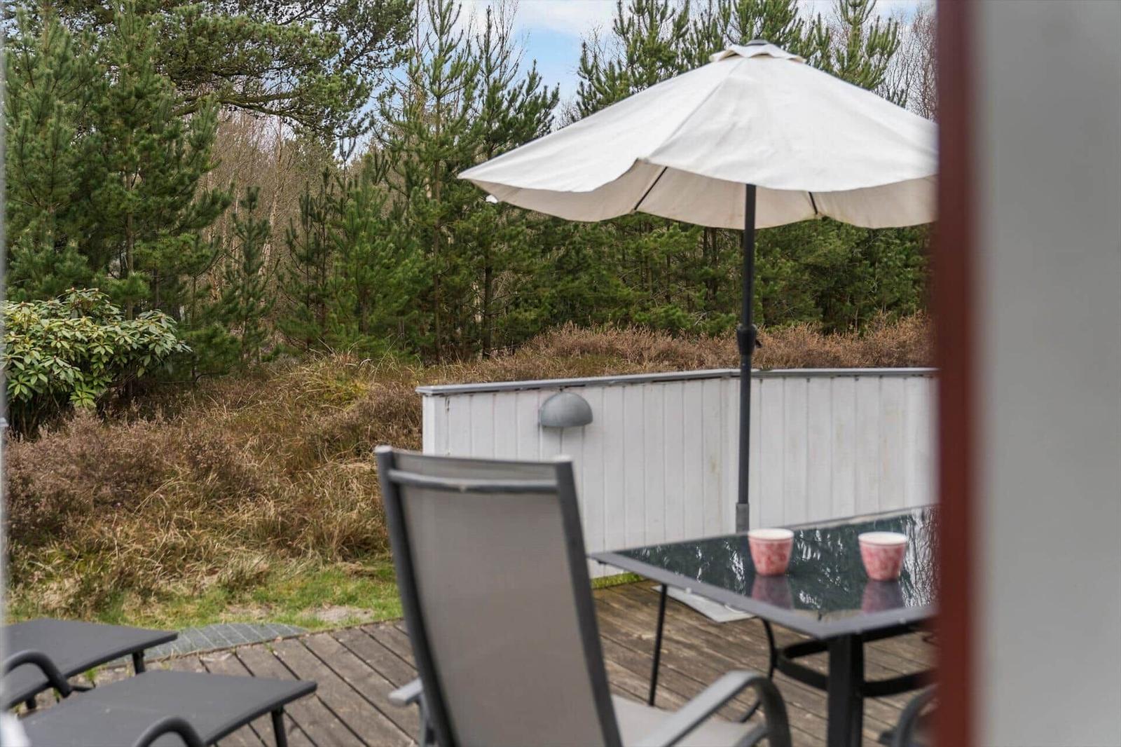 Terrasse mit Tisch, Stühlen und Sonnenschirm. Hintergrund: Wald und Hecke.