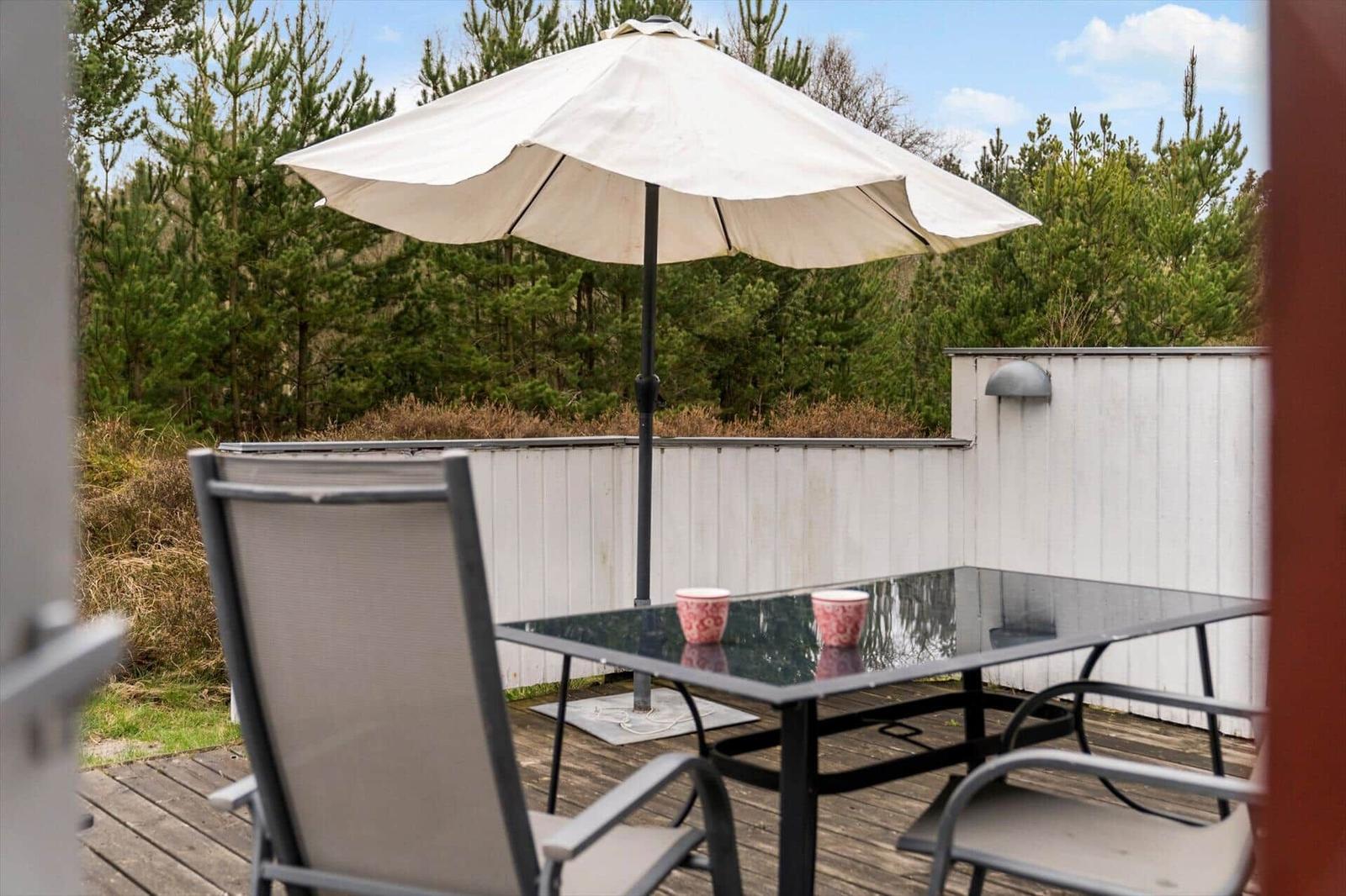 Terrasse mit Tisch, Stühlen und Sonnenschirm. Hintergrund: Wald und weißer Zaun.