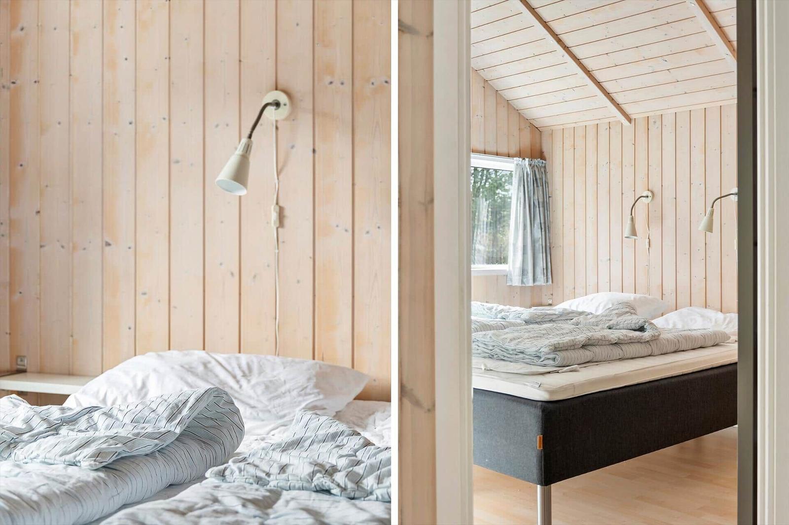 Schlafzimmer mit Holzwand und Bett. Fenster mit Vorhang. Deckenleuchten an der Wand.