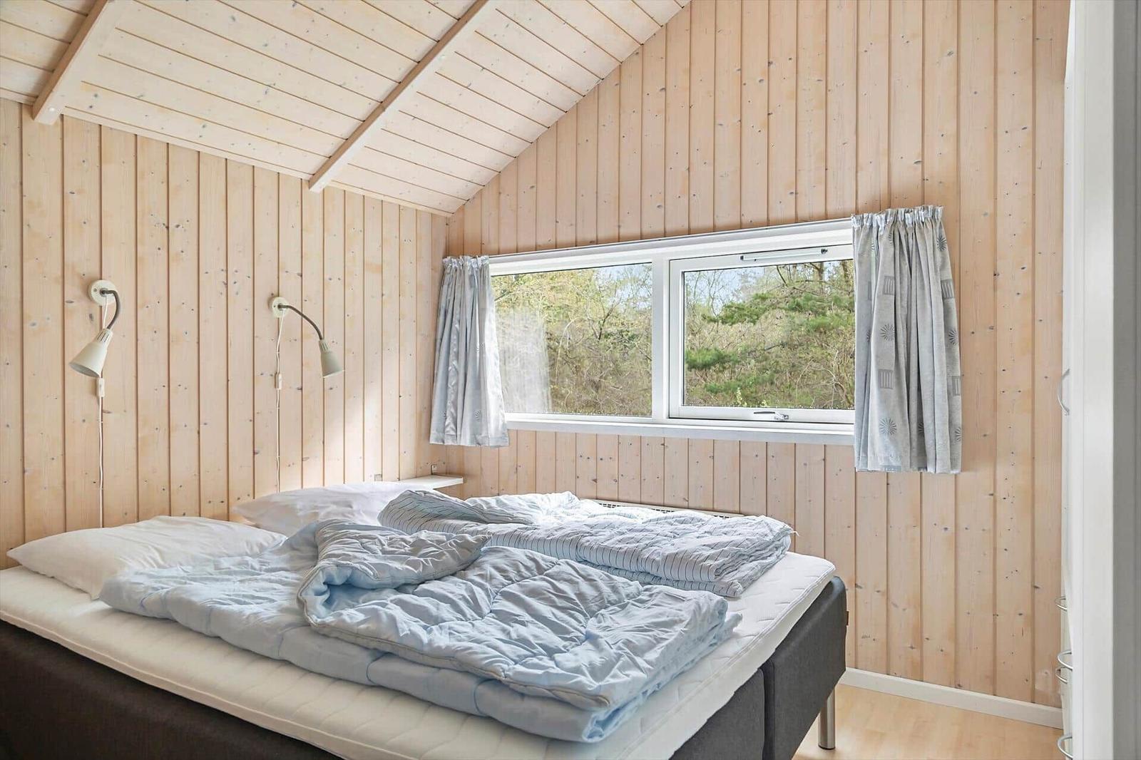 Schlafzimmer mit Holzwand und Dachfenster. Bett mit blauer Bettwäsche und Vorhängen.