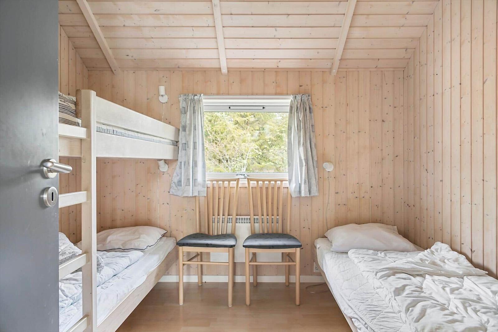 Zimmer mit Doppelstockbett, Einzelbett und zwei Stühlen. Holzwände und Fenster mit Vorhängen.