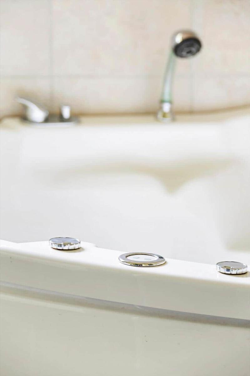 Eine weiße Badewanne mit drei Chromknöpfen und einem Duschbrausekopf im Hintergrund.