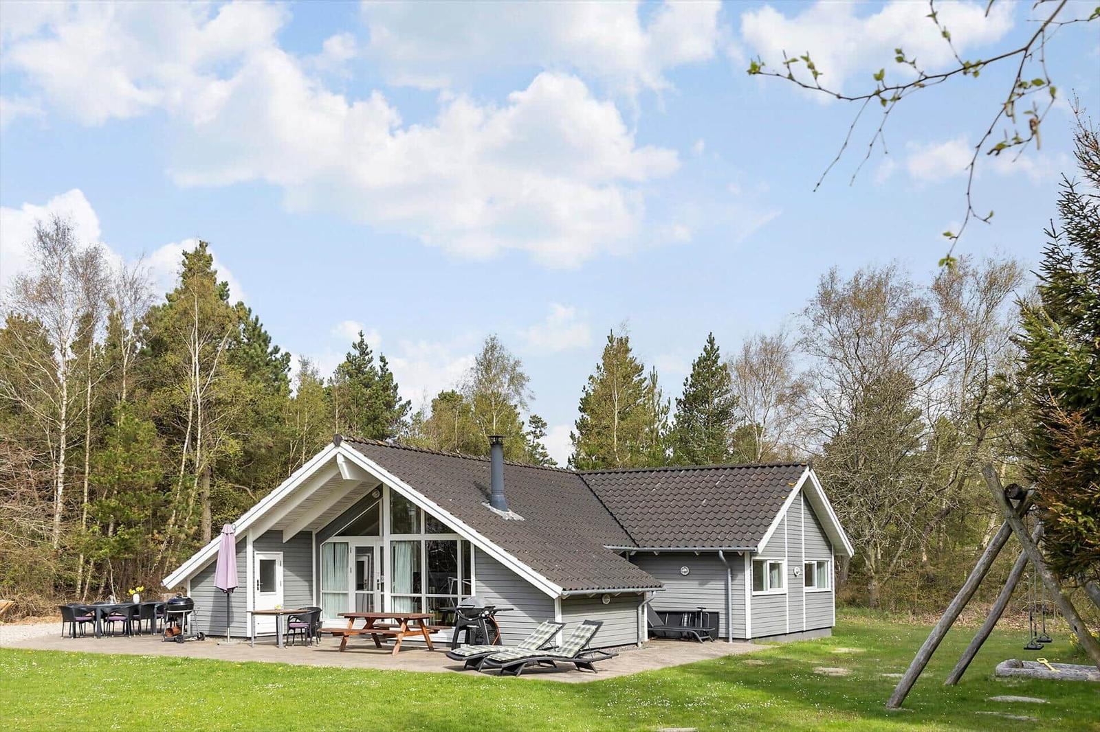 Ein graues Ferienhaus mit Terrasse, Grill und Liegen im Grünen.