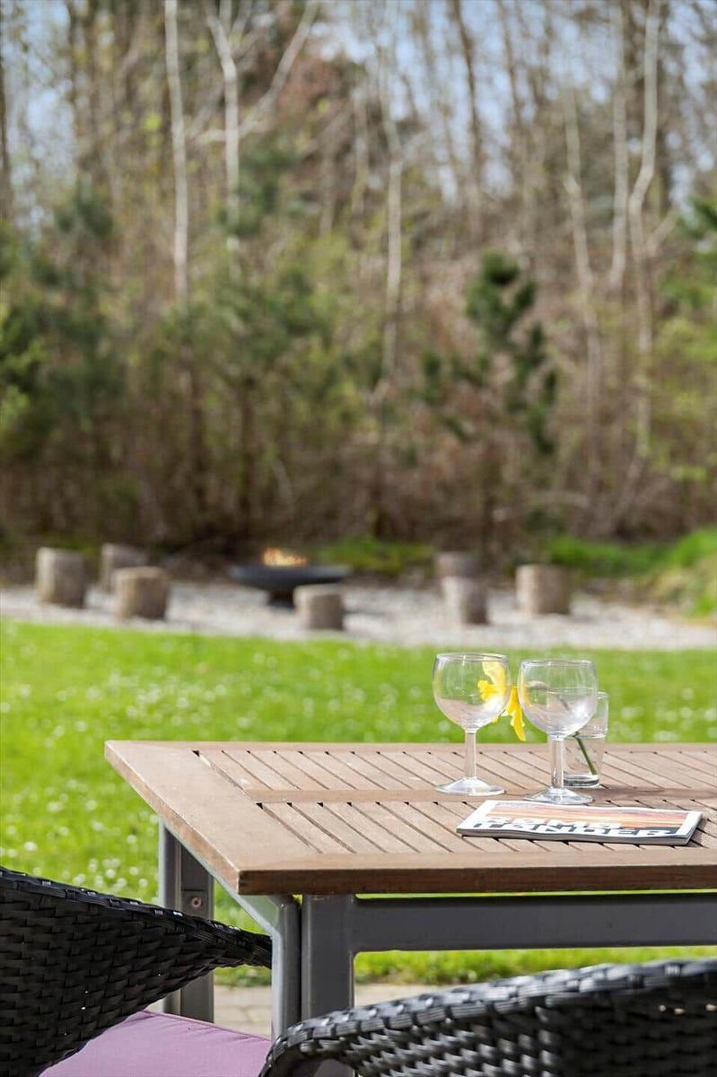 Holztisch mit zwei Gläsern und Zeitschrift im Garten mit Feuerkorb und Grünfläche.