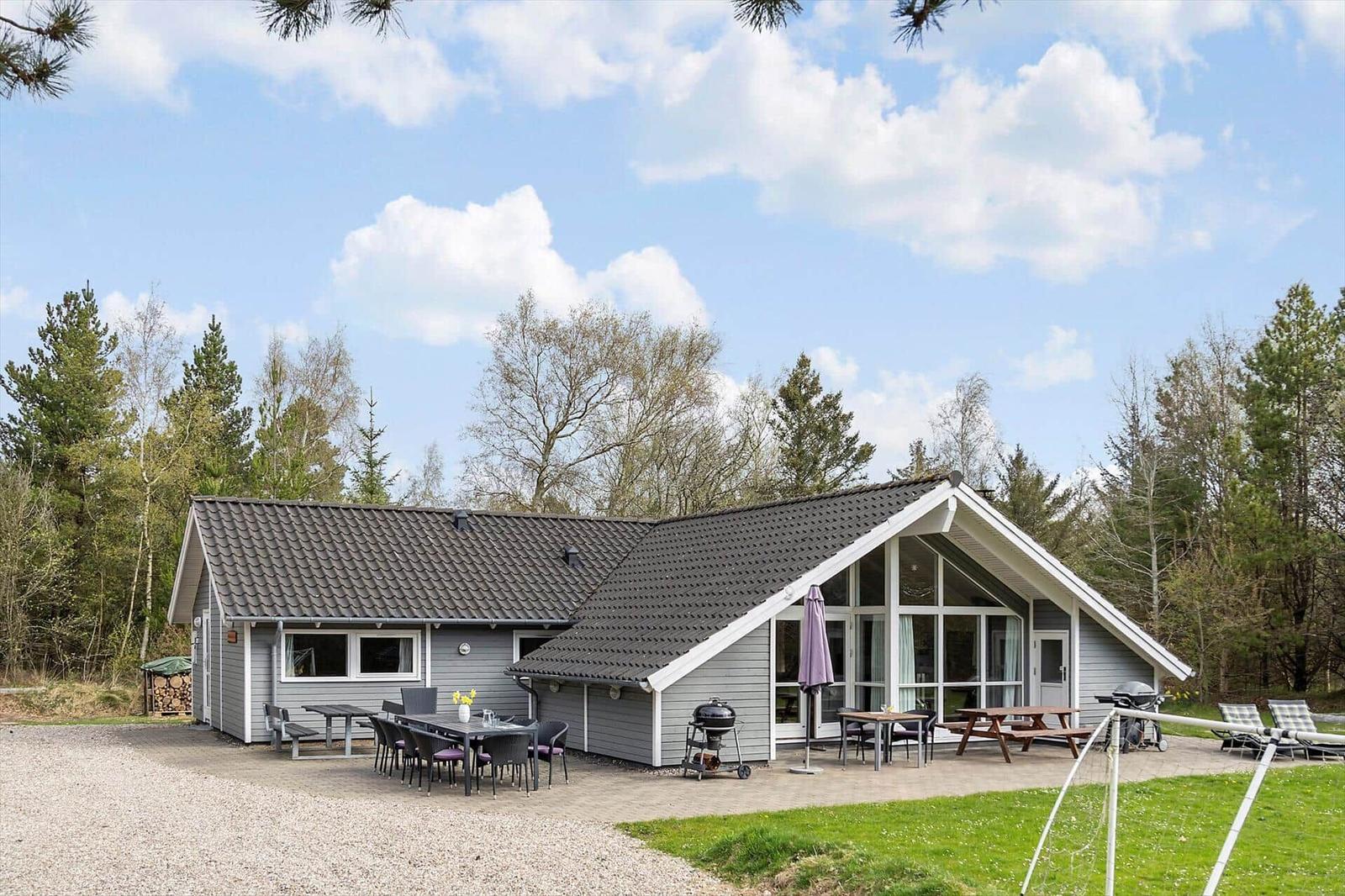 Ein Ferienhaus mit Terrasse, Grill und Garten. Umgeben von Bäumen unter blauem Himmel.