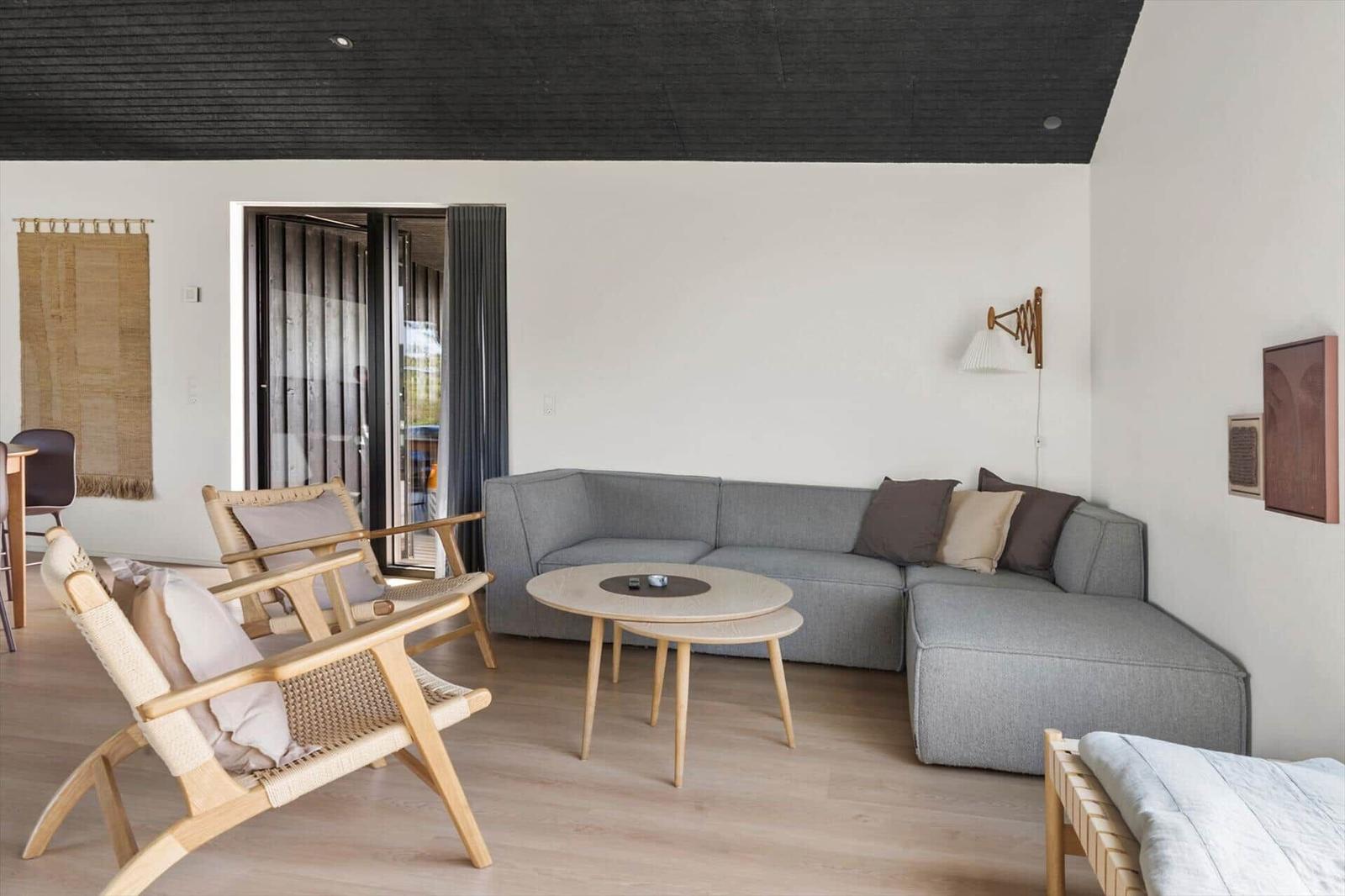 Wohnbereich mit grauem Sofa, Holzstühlen und runden Tischen. Schwarzer Deckenrahmen und weiße Wände.