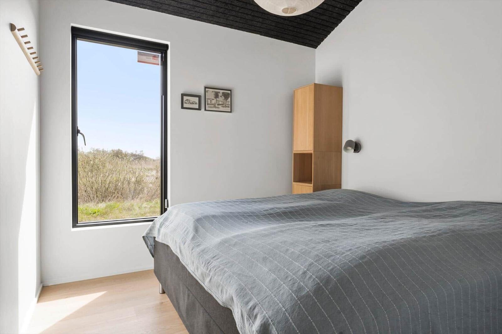 Schlafzimmer mit Bett, Fenster und Holzregal. Blick auf Grünflächen.