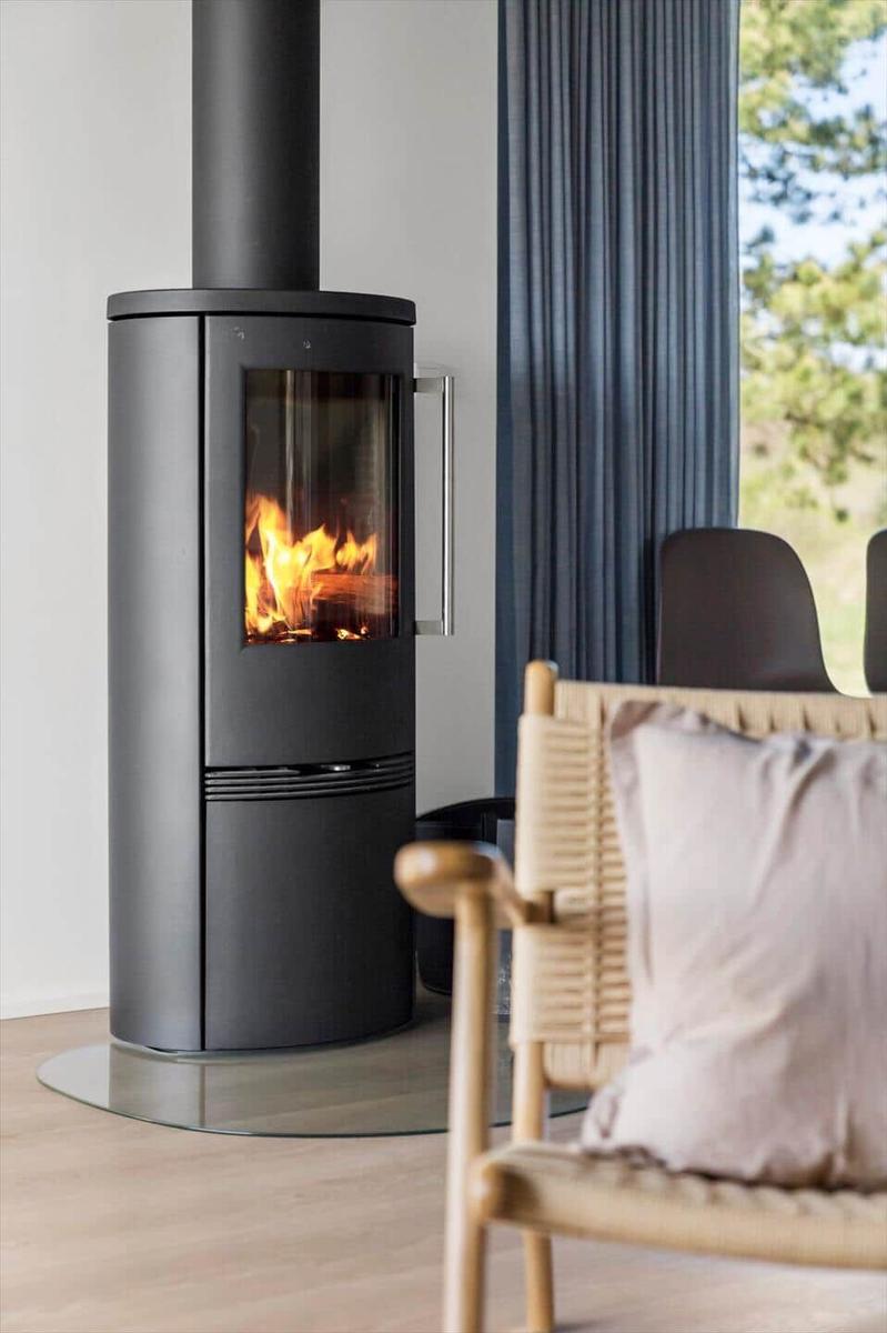 Moderne Feuerstelle mit brennendem Holz, daneben Stuhl mit Kissen und Fenster mit Vorhang.